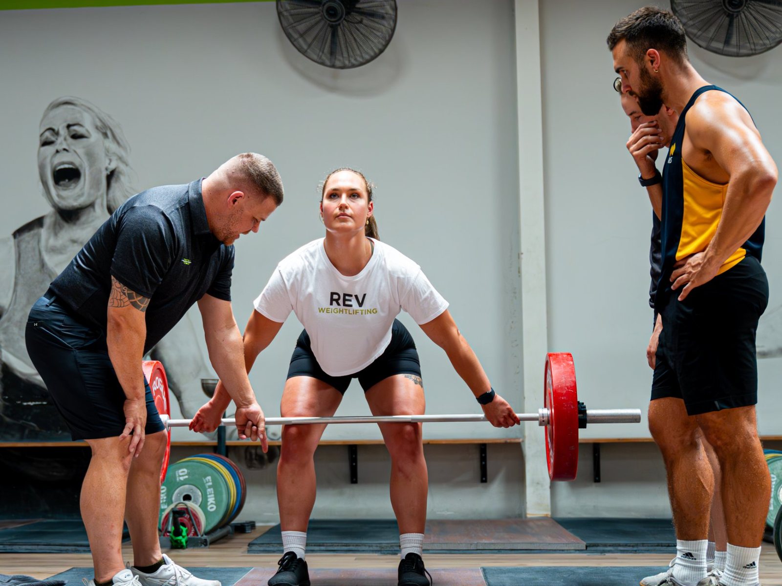 A coach checks a lifter's starting position at the bar before a snatch