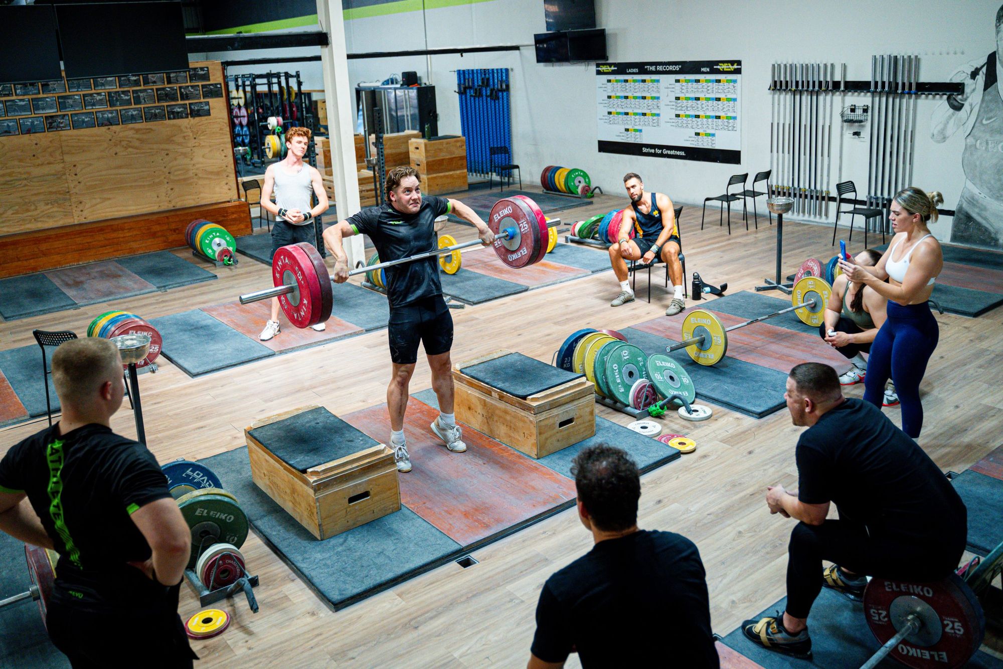 A lifter at Rev Barbell Club pulls a heavy barbell from blocks while teammates and coaches watch